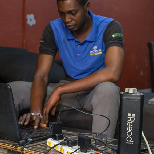 A young man types on a laptop that is plugged into a milk-carton-sized battery pack. The name Reeddi is written on the pack.