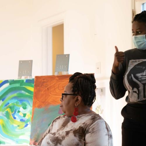 On a film set depicting an art studio, Nana Frimpong, wearing a mask, gives the thumbs up to actors.