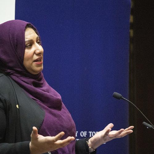Ausma Malik holds both hands palms up as she gestures while speaking at a podium.