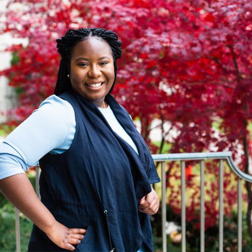 Nicole Mfoafo-M'Carthy smiles as she stands hand on hip in a garden outdoors.