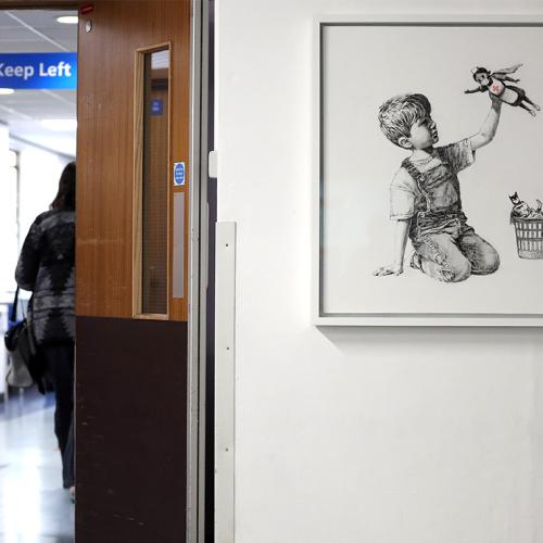 A nurse walks past a hospital artwork showing a small boy playing with 3 superhero toys: batman, spiderman and a nurse.