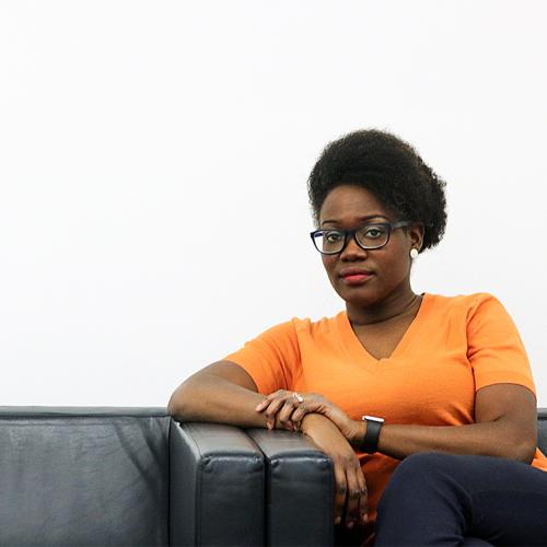 Onye Nnorom looks serious, sitting alone on a sofa with her elbow on one of its arms.