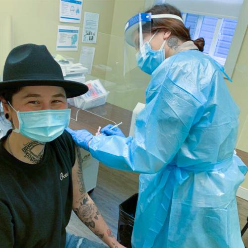 A health-care worker in protective equipment gives a vaccination to a smiling patient in a mask.
