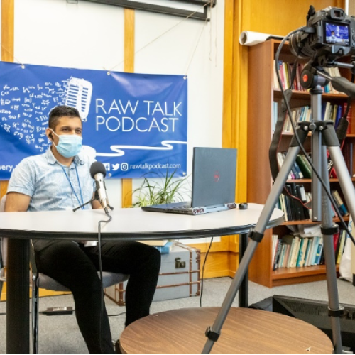 Yagnesh Ladumor, wearing a mask, sits at a table with a large microphone, in front of a banner that reads Raw Talk Podcast.