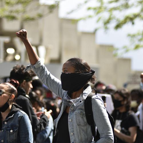 A young woman in a mask holds her fist in the air. Behind her is a crowd of other protesters on University Avenue in Toronto.
