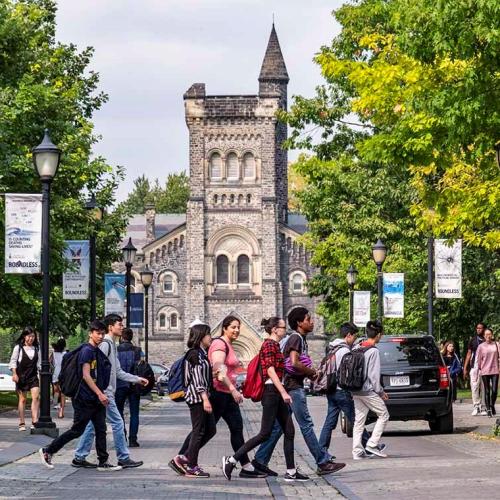 Students wearing backpacks cross the road. University College peeks through trees in the background.