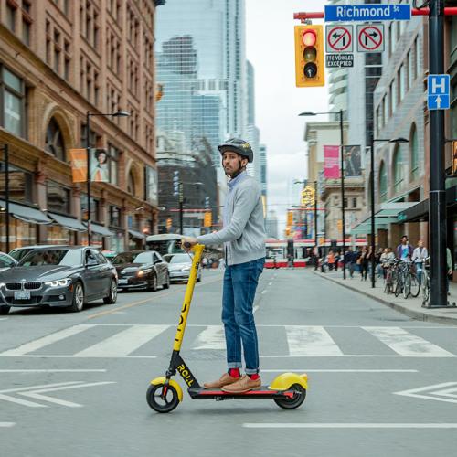 A young man wearing a helmet stands on an e-scooter as he crosses College Street in downtown Toronto.