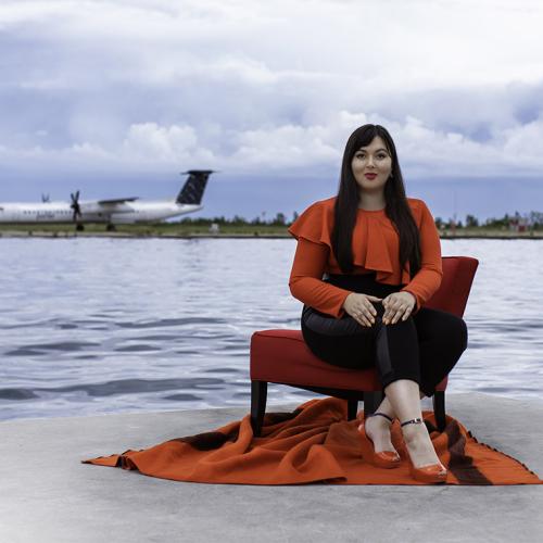Lesley Hampton smiles as she poses in a dramatic ruffled shirt, on a dock across from a lakeside airport.