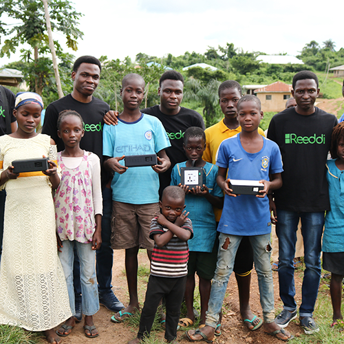 Four men wearing Reedi T-shirts pose with a group of children holding Reedi devices in a village among trees.