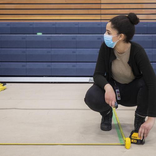 Reem El-Ajou, wearing a mask, uses a tape measure to mark off a social distancing line on the floor of a gymnasium.