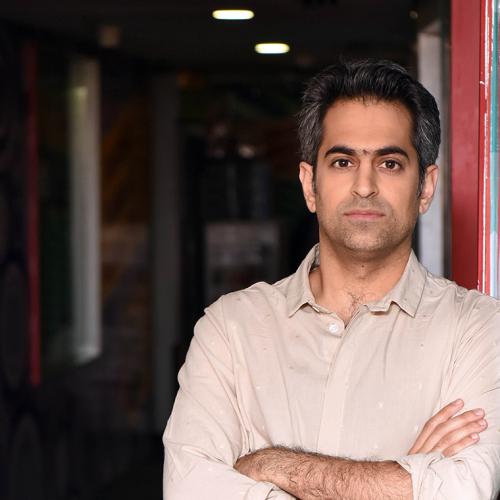 Richie Mehta looks serious as he leans on a glass wall indoors with arms folded.