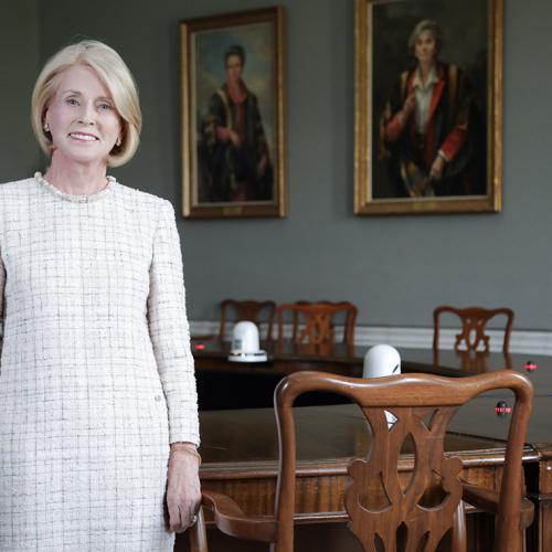 Rose Patten smiles as she stands in front of a large boardroom table, in a room with painted portraits on the wall.