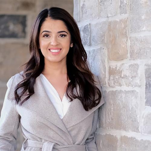 Rumeet Billan smiles as she stands outdoors by a stone wall.