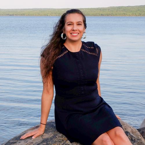 Shanna Peltier smiles, sitting on a large rock on the shores of a lake.