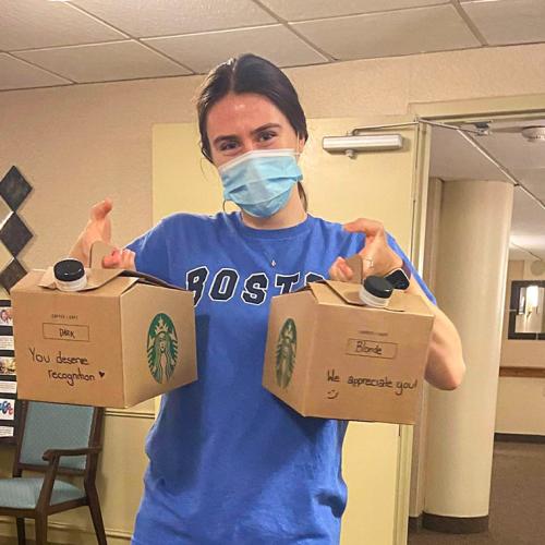 A woman in a mask holds up cardboard boxes of coffee with the words You deserve recognition written on the side.