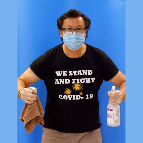 Stephen Lew wears a mask and a T-shirt reading We Stand and Fight COVID-19. He holds cleaning supplies in his gloved hands.