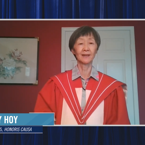 Shirley Hoy, wearing academic robes, speaks on video while standing in front of a painting of flowers.