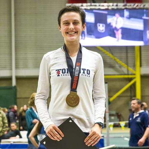 Gabriela DeBues-Stafford smiles, standing in a gym wearing a U of T shirt and a gold Track and Field 2017 medal.