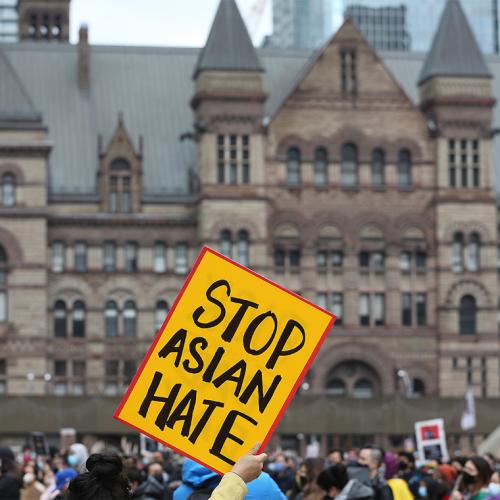 A handlettered sign reading Stop Asian Hate is held high above a crowd in front of Toronto's Old City Hall.