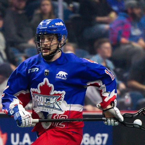 Mitch De Snoo, in the Rock lacrosse team uniform, runs with the ball in his net while a crowd cheers.