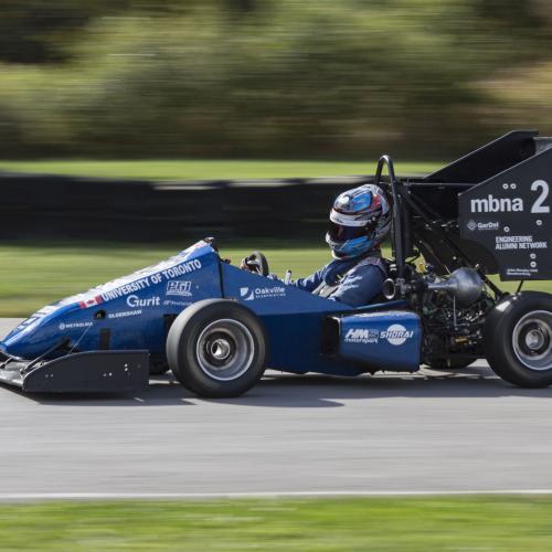 The half-sized U of T Formula SAE racing car speeds down a track. It features an open cockpit, rollbar, and huge rear wing.