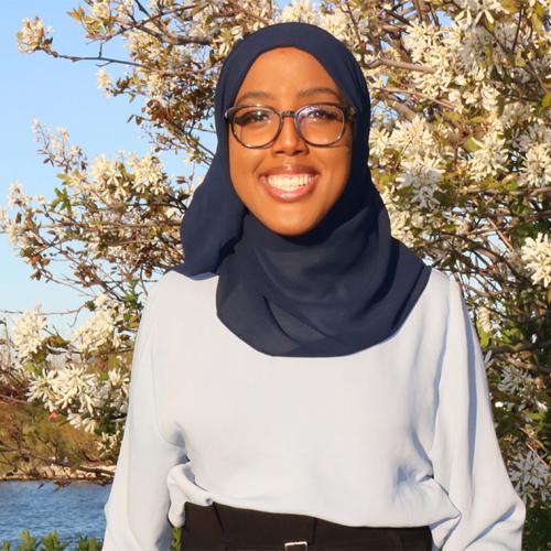 Habon Ali smiles happily, standing by a flowering shrub next to a small lake.