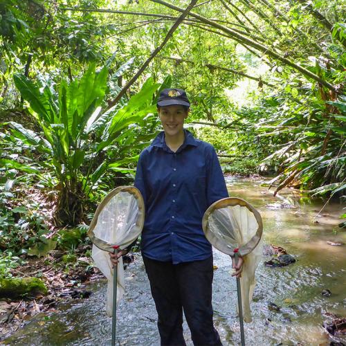 Alex De Serrano stands in a shallow stream, holding two scoop-shaped nets. Lush vegetation arches overhead.