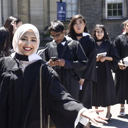 A woman smiles and gestures while standing in a line of diverse students in academic robes waiting to graduate.
