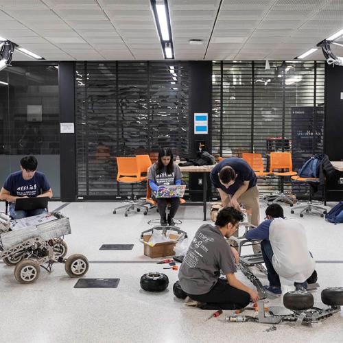 Five students crouch intently over their projects scattered across a classroom floor--a wheeled square object, a frame on tank tracks, and a drone.