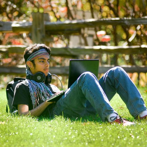 A young man sits alone outdoors on the grass, looking at his laptop screen.