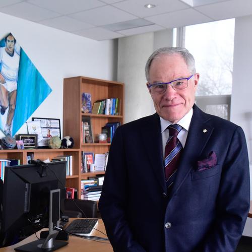 Bruce Kidd smiles as he stands in his office. On the wall are three Olympic torches and a painting of a young Kidd running.