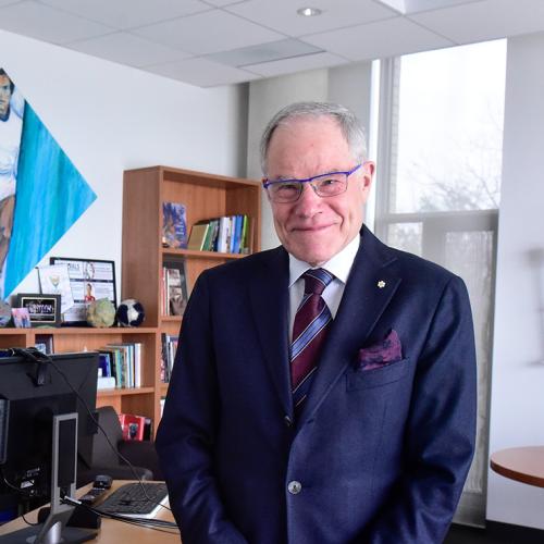 Bruce Kidd smiles as he stands in his office. On the wall are three Olympic torches and a painting of a young Kidd running.