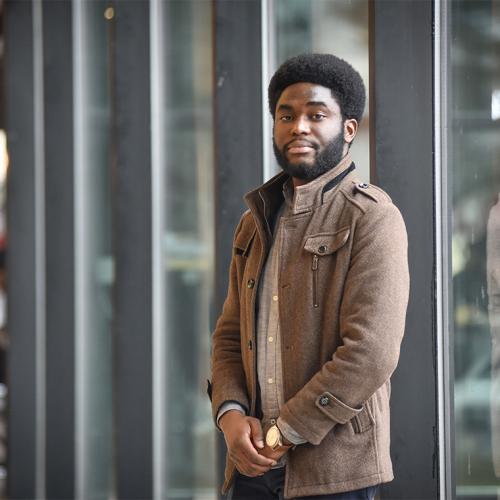 Efosa Obano smiles as he stands outside in front of a glass-walled building.