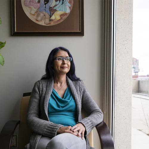 Julie Blair looks thoughtful as she sits by a large window near a painting in an Indigenous style.