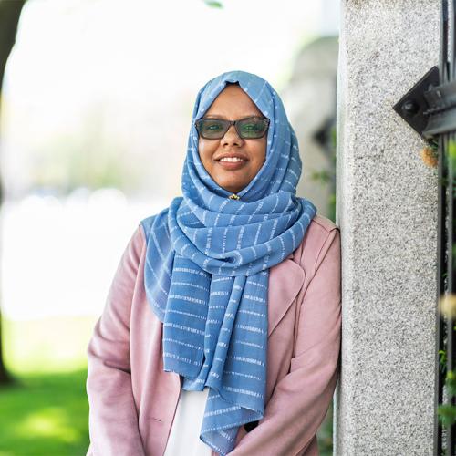 Mariam Abeid smiles as she leans on an iron fence near a tree outdoors.