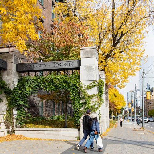 A couple walk out of the Alumni Gates on College Street, beneath the sign reading University of Toronto.