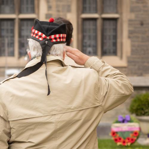 An older man, wearing a ceremonial military cap, salutes a row of wreaths.