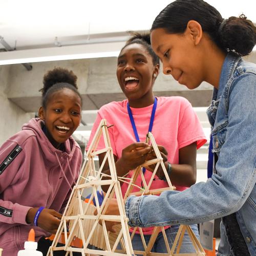 Three young girls laugh with delight as they construct a model bridge out of popsicle sticks.