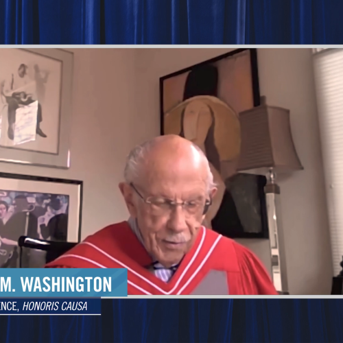 Warren Washington, wearing academic robes, speaks on a video call from a room decorated with paintings.