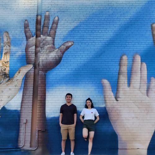 Weiwei Li and Catherine Chan smile as they pose beside a wall mural featuring several giant raised hands.