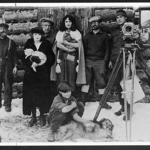 In 1923, director Nell Shipman stands in snow in front of a log cabin, with a film camera and several crew and cast members.