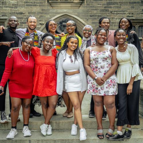 A group of 12 smiling Black graduates and their friends and family stand outside of Hart House.