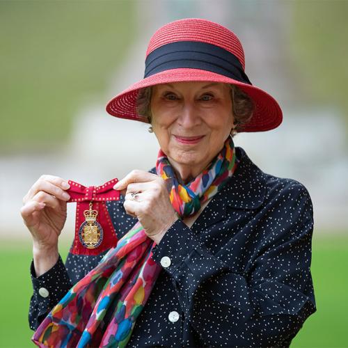 Margaret Atwood smiles and holds up a medal topped with a crown and mounted on a ribbon tied in a bow.