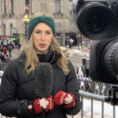 Annie Bergeron-Oliver holds a microphone and speaks to a camera in Ottawa, with protesting trucks behind her.