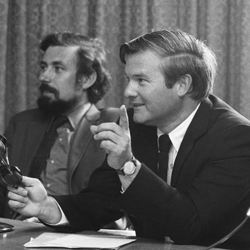 In a photograph from the 1970s, Bill Davis leans on a table and speaks into a microphone.