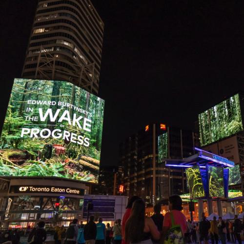 People in a plaza at night look up at a screen. Text reads, Edward Burtynsky's In the Wake of Progress.