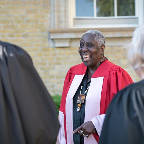 Camille Orridge, wearing academic robes, laughs out loud as she chats with other academics in robes.