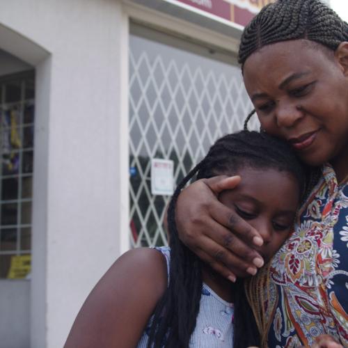 In a still from the film Because She Cares, a woman hugs a little girl who is leaning on her.