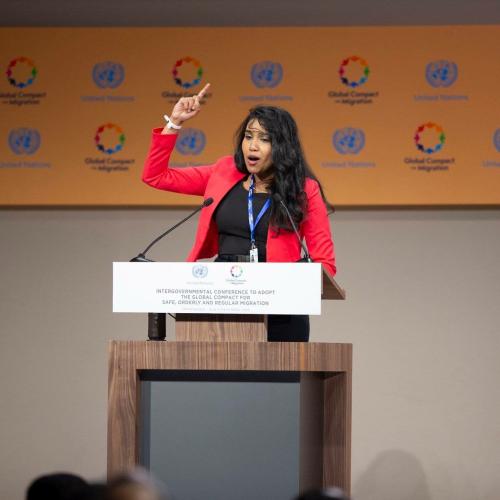 A woman speaking at a podium and gesturing with her right arm. 