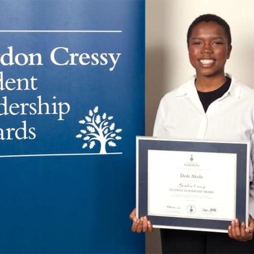 Dede Akolo smiles and holds a certificate next to a sign reading: Gordon Cressy Student Leadership Awards.